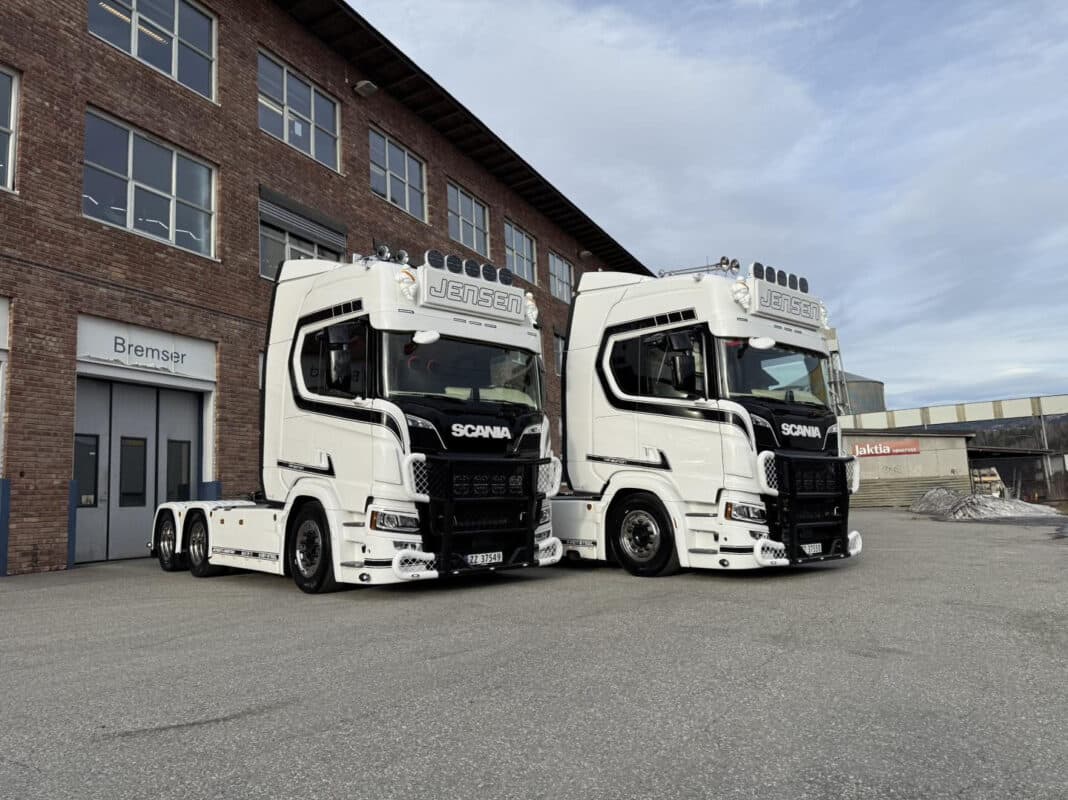 To hvite Scania-lastebiler med “Jensen” på toppen parkert foran et industribygg.