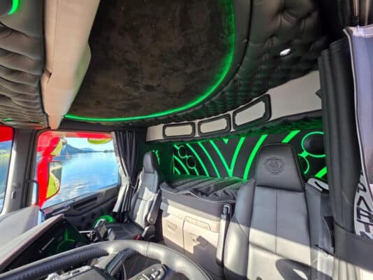 Interiør i lastebil med svarte skinnseter og grønn LED-belysning, sett mot vann og fjell utenfor vinduet.